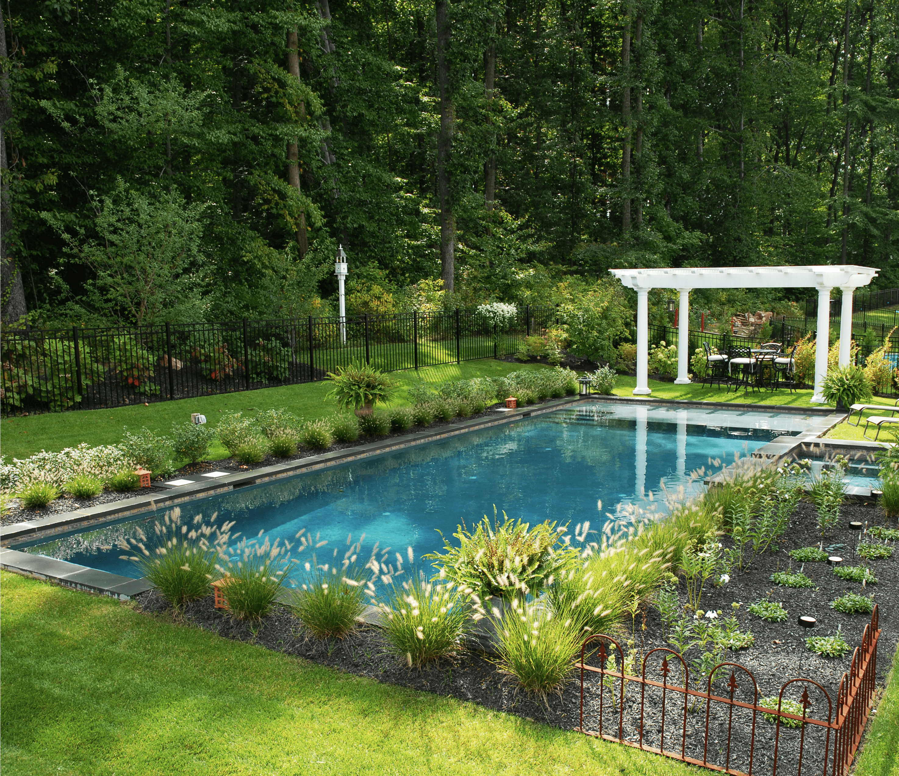 A professionally landscaped backyard featuring a rectangular swimming pool with an integrated spa. The pool area is surrounded by lush green lawn, various plants, and a small decorative fence in the foreground. A white pergola with outdoor dining furniture is positioned at the far end of the pool.