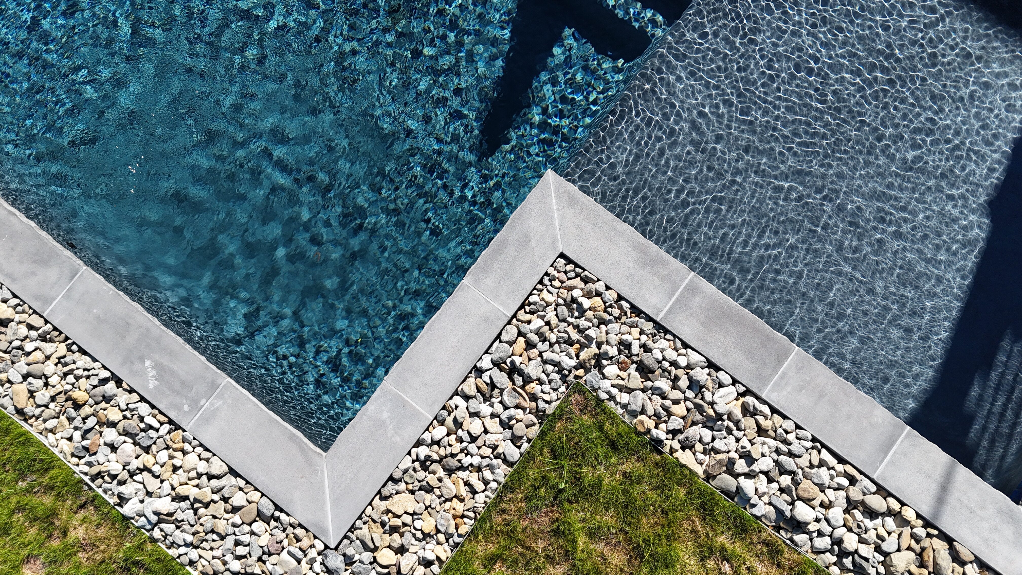 An overhead shot of the corner of a swimming pool. The pool's coping is a dark gray stone, creating a sharp angle. The pool has deep blue water. The deck area is a mix of small, light-colored river stones and a patch of green grass, creating a modern and natural look.