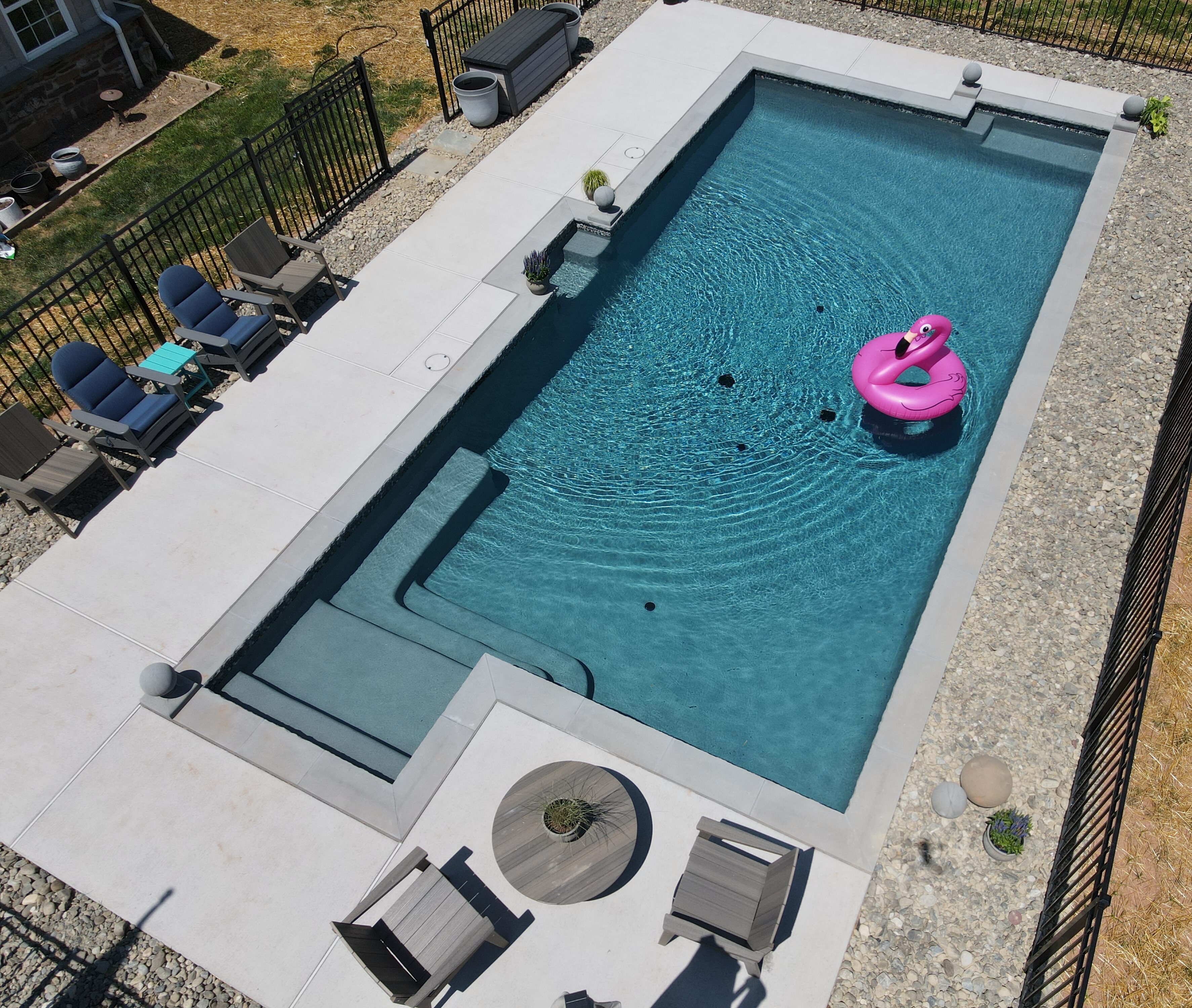 An aerial view of a rectangular, modern swimming pool with a deep blue interior. The pool has integrated steps and a bench. A pink flamingo float is in the water. The pool is surrounded by a light-colored concrete patio and a border of small gray stones. The patio has a few lounge chairs and a small dining set. A black metal fence encloses the backyard.