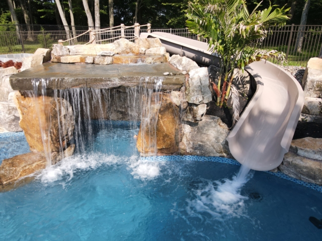 A close-up view of a custom swimming pool with a large, elaborate waterfall and a slide. The waterfall is made of natural-looking stone and a flat rock, with water cascading into the pool. To the right of the waterfall, a tan-colored, curved pool slide descends into the water.