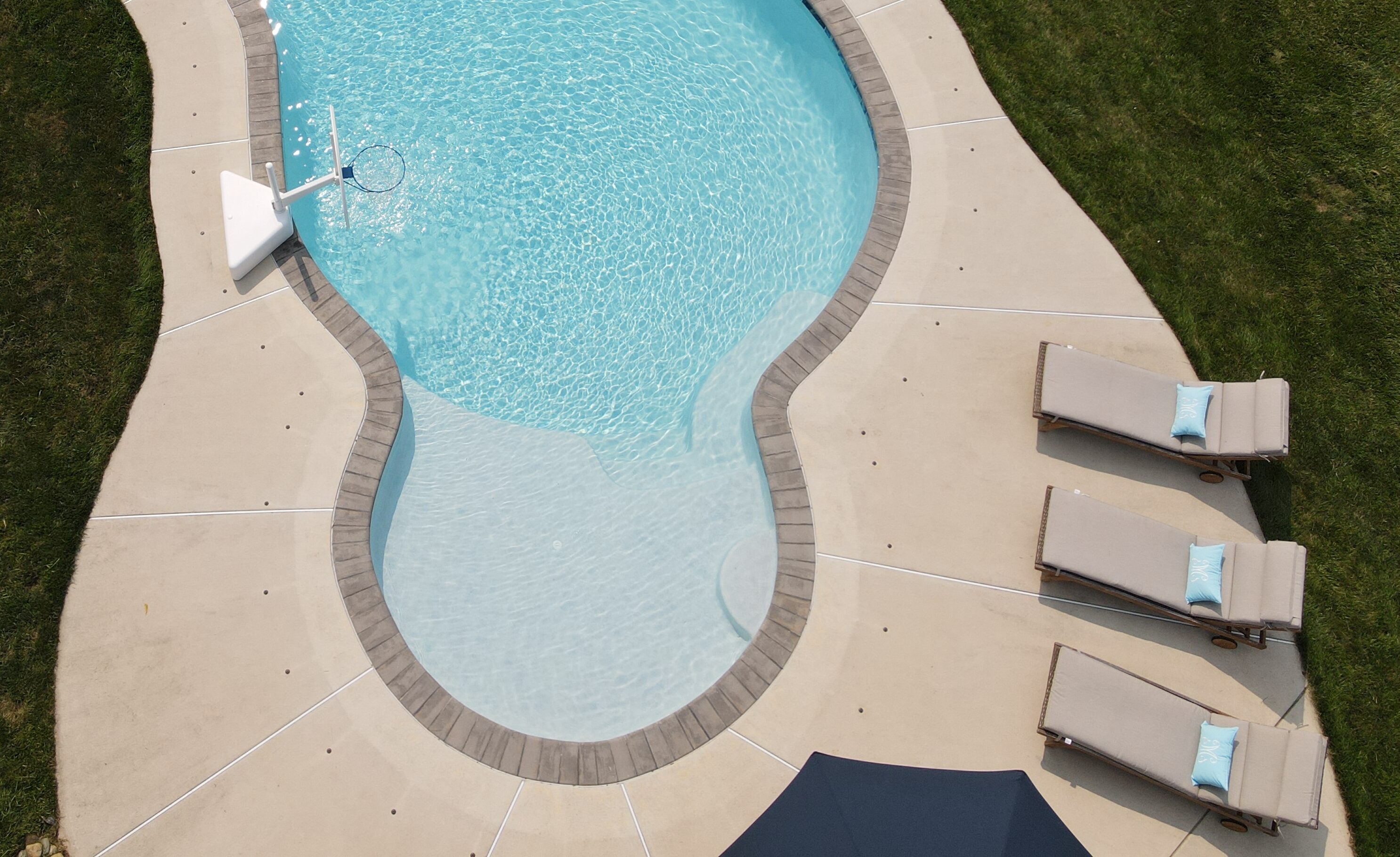 An aerial shot shows a curved, kidney-shaped swimming pool with dark tan coping. The pool has a basketball hoop on one end and is surrounded by a light-colored concrete patio. On the right side of the patio, three identical lounge chairs with light blue pillows are arranged in a row. A dark blue umbrella is visible at the bottom of the frame, near the chairs. Beyond the patio is a green lawn.