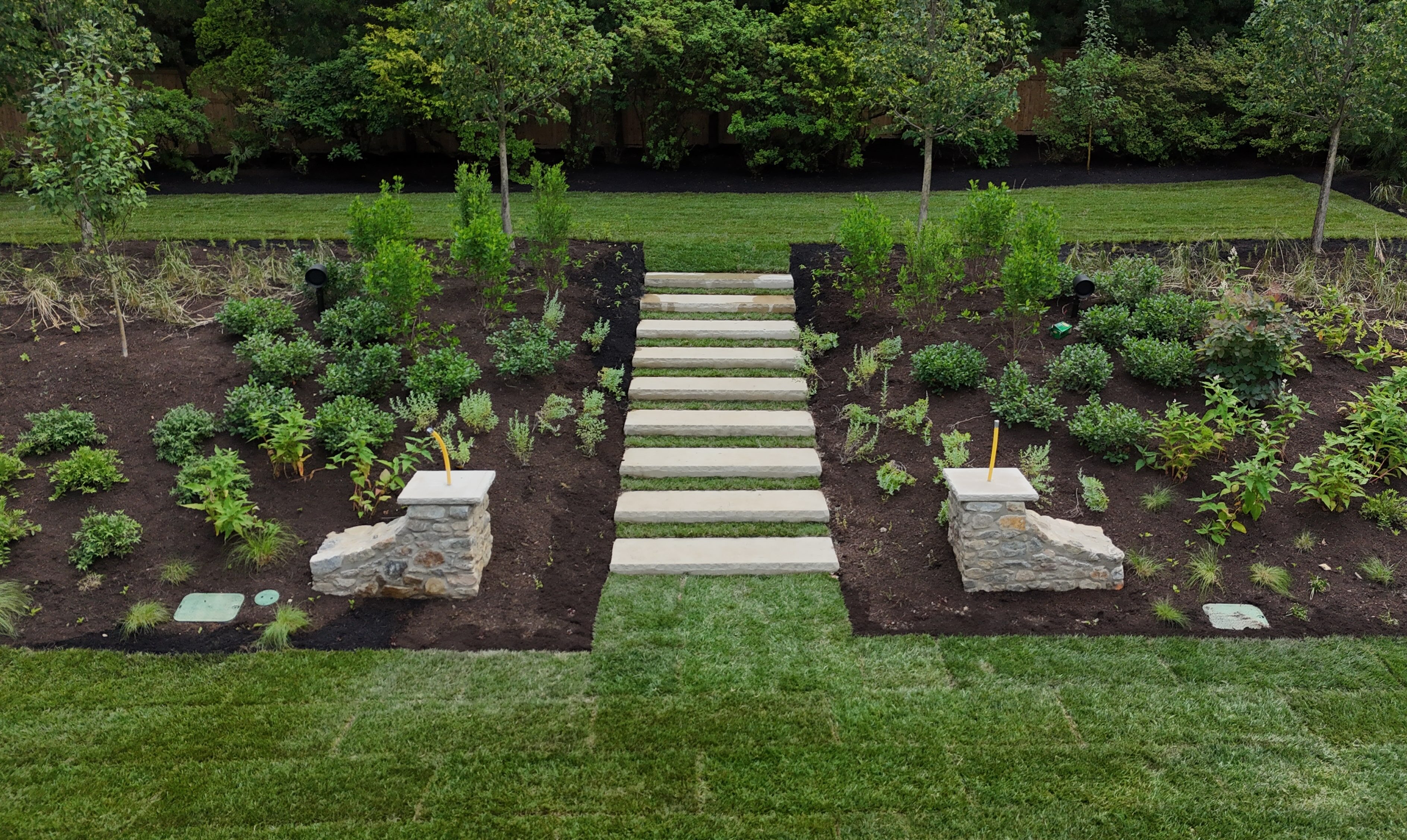 A beautifully landscaped backyard with a set of modern stone slab steps leading up a gentle slope. The steps are flanked by stone pillars and surrounded by garden beds filled with dark mulch and young plants. The area is bordered by a well-maintained green lawn at the bottom and a row of trees and bushes at the top.