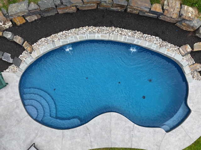 An aerial shot of a unique, kidney-shaped swimming pool. The pool is surrounded by a light-colored concrete patio. The far edge of the pool has a stone coping and a bed of white pebbles. The pool itself is a deep blue, with steps at one end and a bench seat at the other.