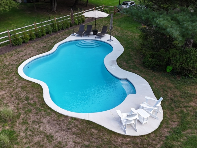 An aerial shot of a backyard with a large, irregularly shaped swimming pool. The pool has light blue water and is surrounded by a white concrete patio.