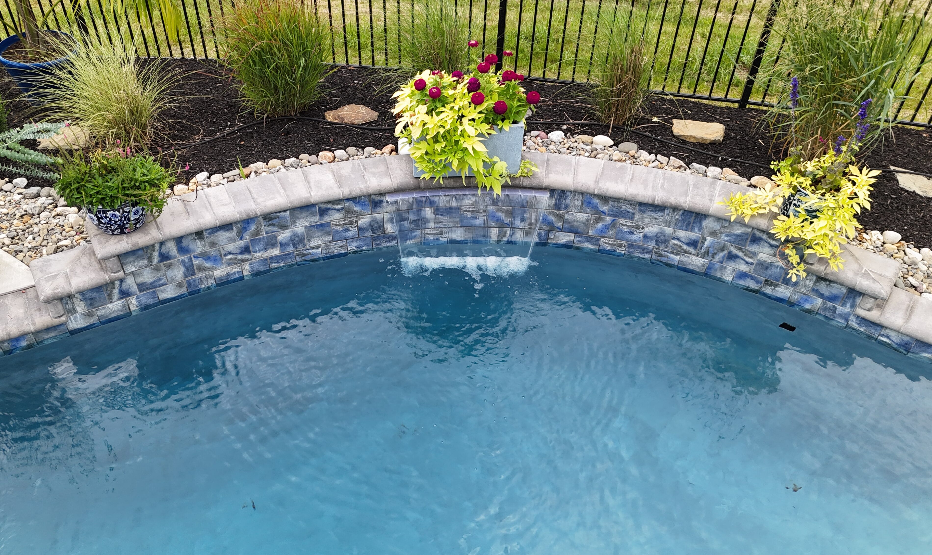 A backyard swimming pool with a stone wall and small waterfall feature. The wall is made of textured, blue-and-gray tiles and is topped with light gray coping stones. Three decorative pots with plants sit on the coping stones, one on the left, one in the middle, and one on the right. The area behind the wall is landscaped with mulch, river rocks, grasses, and other plants. A black metal fence runs along the back of the property.