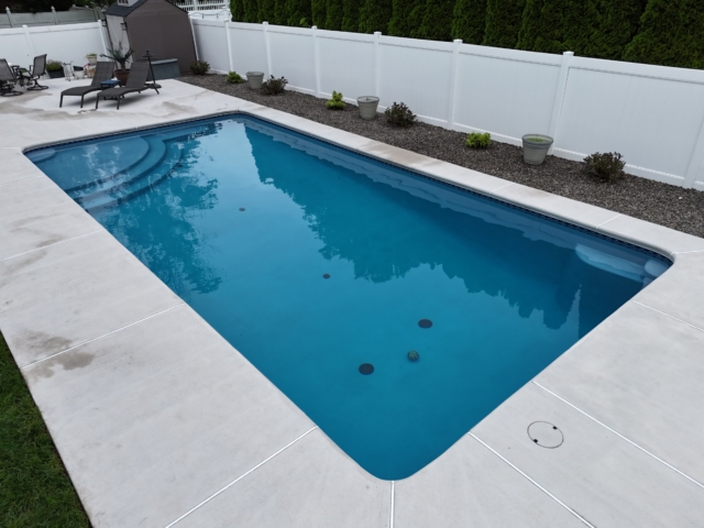 A ground-level shot of a rectangular swimming pool with a light blue interior. The pool has rounded corners and is surrounded by a wide, gray concrete patio. At the far end of the pool, a section with built-in steps is visible.