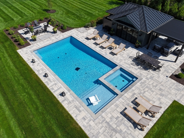 An aerial view of a luxurious, modern backyard with a rectangular swimming pool and a square hot tub. The pool features an integrated shallow sun shelf with a submerged white lounge chair. Surrounding the pool is an expansive patio made of light-colored pavers. A dark-roofed pool house with a matching pergola is located next to the pool.