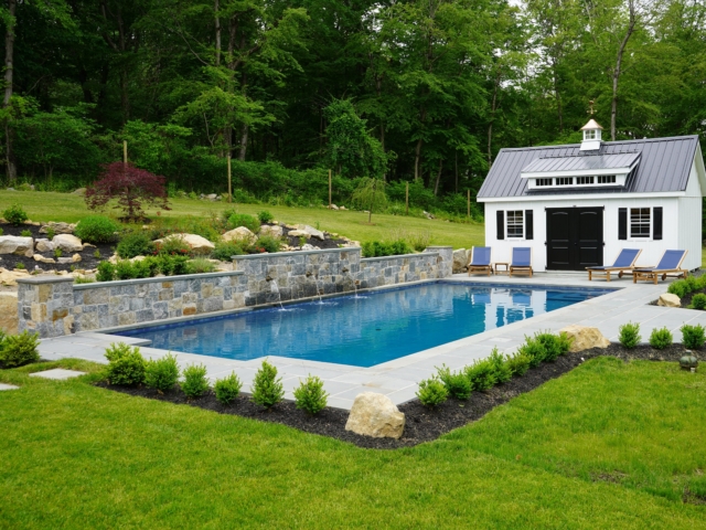 A rectangular swimming pool in a beautifully landscaped backyard. The pool has a natural stone wall at its far end with three small waterfalls spilling into the water. Surrounding the pool is a gray paver patio. A white shed with a dark gray metal roof and black doors and shutters stands next to the pool.