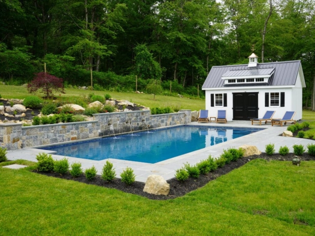 A rectangular swimming pool in a beautifully landscaped backyard. The pool has a natural stone wall at its far end with three small waterfalls spilling into the water. Surrounding the pool is a gray paver patio with several blue lounge chairs. A white shed with a dark gray metal roof and black doors and shutters stands next to the pool.