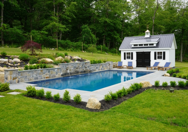 A rectangular swimming pool in a beautifully landscaped backyard. The pool has a natural stone wall at its far end with three small waterfalls spilling into the water. Surrounding the pool is a gray paver patio with several blue lounge chairs. A white shed with a dark gray metal roof and black doors and shutters stands next to the pool.