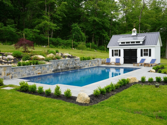 A rectangular swimming pool in a beautifully landscaped backyard. The pool has a natural stone wall at its far end with three small waterfalls spilling into the water. Surrounding the pool is a gray paver patio with several blue lounge chairs. A white shed with a dark gray metal roof and black doors and shutters stands next to the pool.