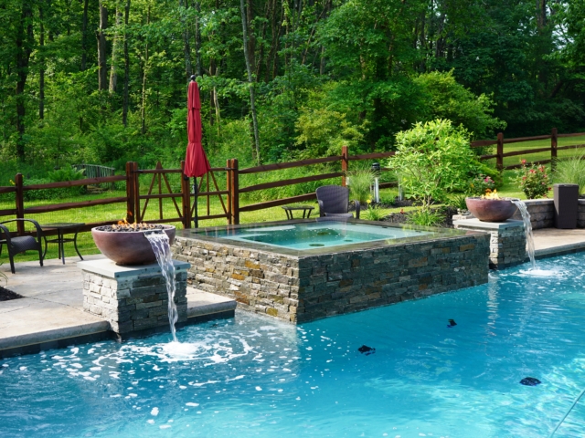 A beautiful swimming pool with a raised, square hot tub at its center. The hot tub is clad in decorative stone, and two matching stone columns stand at either end. Each column supports a large, bowl-like feature from which water spills into the pool.