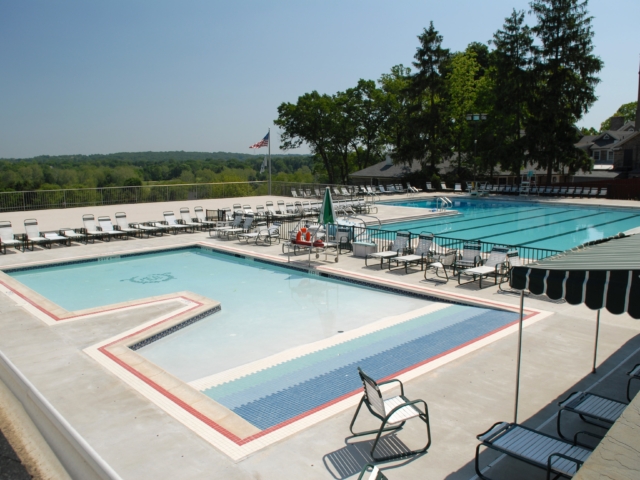 An aerial view of a large, outdoor swimming pool complex with two pools. The larger pool is a rectangular lap pool with blue lane markings. The smaller pool is L-shaped with a shallow area decorated with a mosaic of blue and white tiles. Both pools are surrounded by a concrete deck.