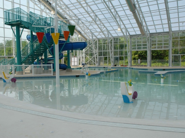 An indoor aquatic center with a large, bright leisure pool and a blue, twisting water slide. The slide structure has a staircase with turquoise metal railings. The building has a high, arched roof with a glass skylight and large windows along the back wall. Two colorful plastic play structures shaped like animals are in the shallow end of the pool.