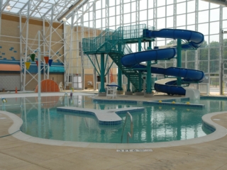 An indoor aquatic center with a large, irregularly shaped leisure pool. The pool features a blue, twisting water slide structure with a metal staircase. In the middle of the pool, there is a small concrete island with blue tile accents. The room has a high, arched ceiling with a large glass skylight and a wall of windows. A small, colorful play structure is visible in the background, along with a lifeguard stand.