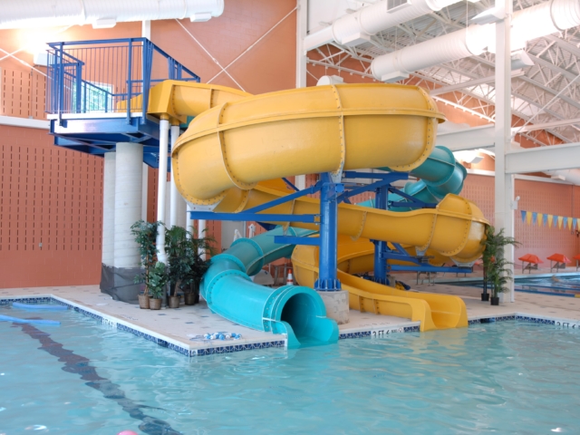 An indoor swimming pool with a large, elaborate water slide structure. The structure has a blue metal frame and features a yellow twisting open slide and a blue enclosed spiral slide. A staircase with blue railings leads to the top of the slides. The pool area has a tiled deck.