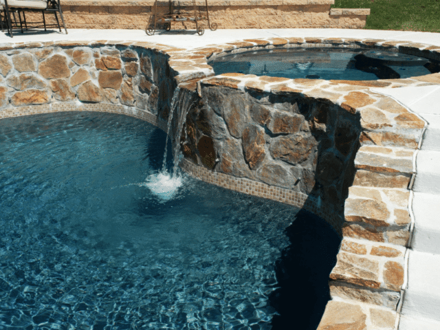 A close-up view of a custom swimming pool and hot tub connected by a stone waterfall. The pool water is a deep blue, and the hot tub is slightly elevated. A curved, rugged stone wall separates the two, with water cascading from the hot tub into the pool.