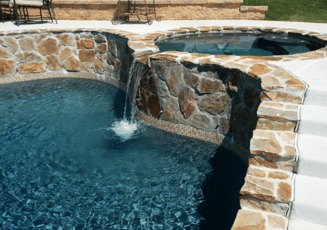 A close-up view of a custom swimming pool and hot tub connected by a stone waterfall. The pool water is a deep blue, and the hot tub is slightly elevated. A curved, rugged stone wall separates the two, with water cascading from the hot tub into the pool.
