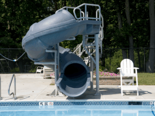 A ground-level view of a swimming pool with a large, dark gray, enclosed spiral water slide. The slide is supported by a white metal frame and stands on the concrete pool deck. To the right of the slide is a white Adirondack chair, and to the left are metal pool handrails.