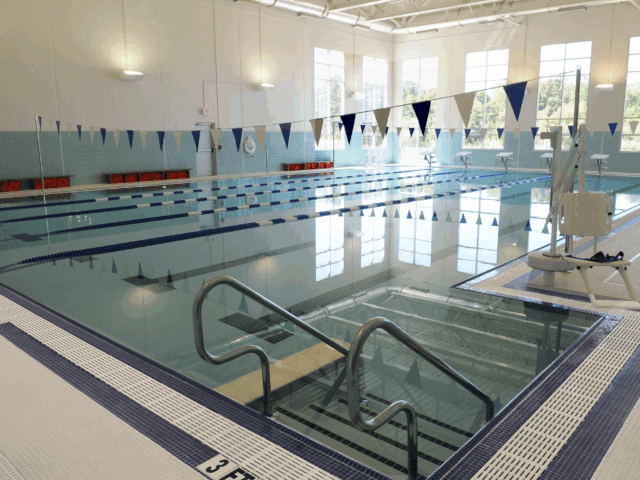 An indoor lap pool with a wide, accessible entry ramp and handrails. The pool has several lanes marked with blue and white dividers. Starting blocks are visible at the far end of the pool.