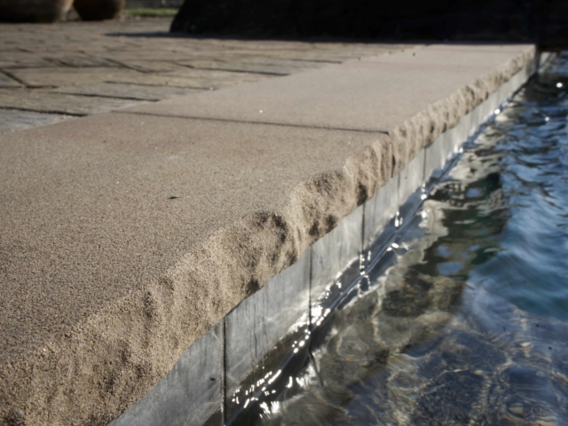 A close-up shot of the edge of a swimming pool. The focus is on the textured, sandy-colored coping stone that meets the water. The water is still and clear, reflecting a ripple of light on the tile below the coping. A brick-patterned patio is visible in the background.