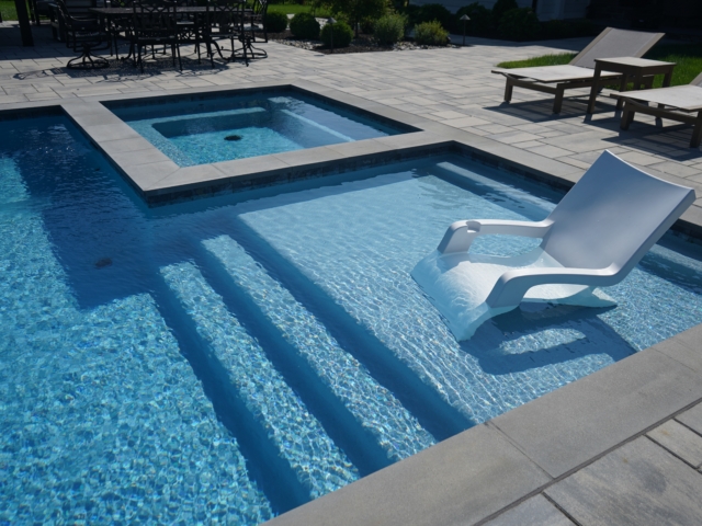 A modern swimming pool with a shallow sun shelf and wide steps leading into the main pool. A white, submerged lounge chair sits on the sun shelf. The pool water is a bright blue. A square hot tub is integrated into the patio next to the pool. The patio is made of light-colored pavers.