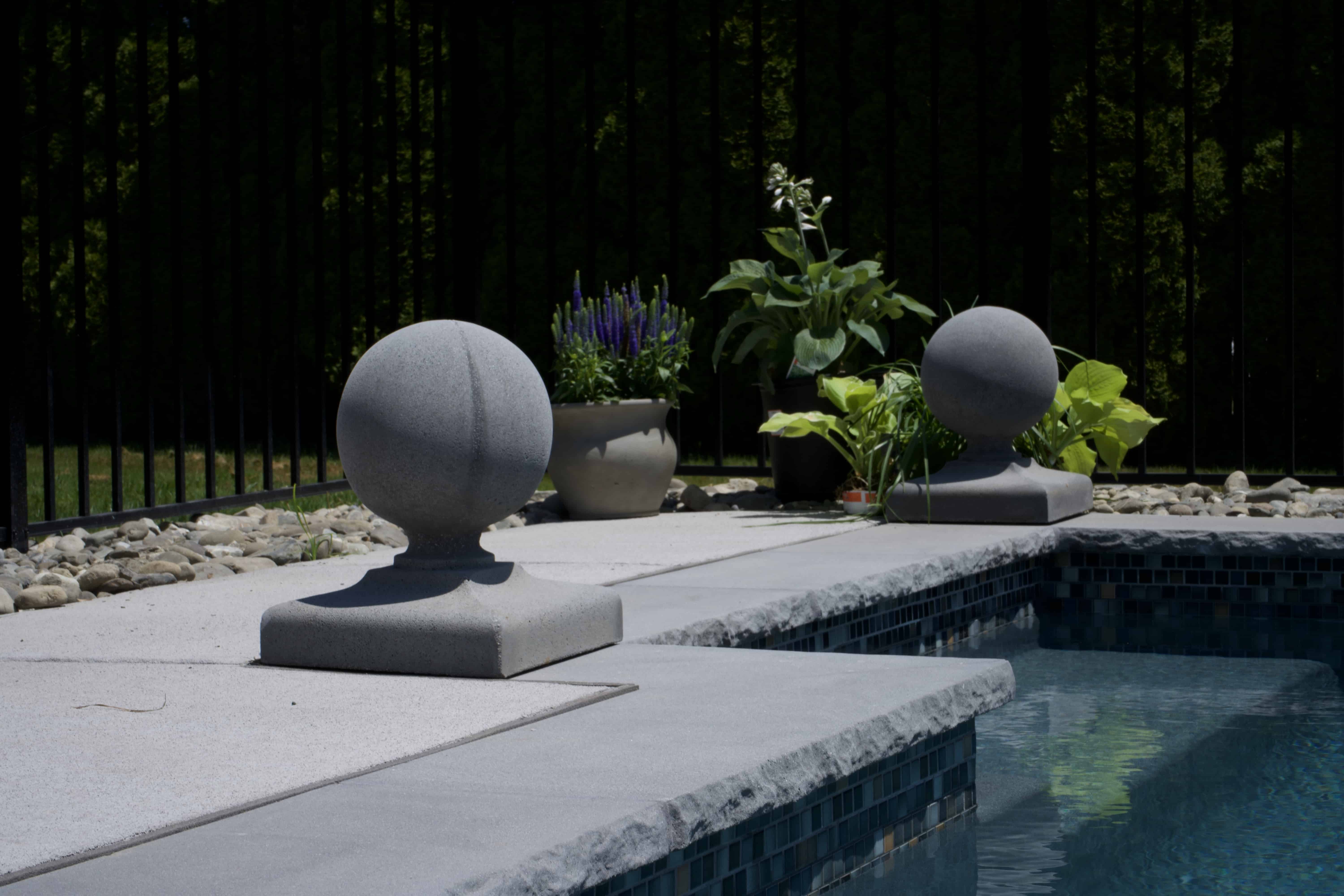 A low-angle shot of a modern swimming pool's edge. The pool coping is a light gray stone with a natural, rough-cut edge. Two decorative gray stone spheres on square bases are placed on the deck, which is made of a lighter-colored concrete. One is in the foreground and the other is further back, near potted plants. The pool water has a mosaic tile border. In the background, there is a black metal fence and a line of dark green shrubs.