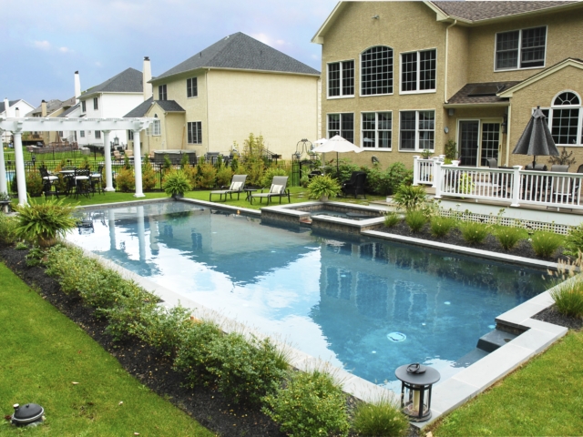 A long, rectangular swimming pool with an integrated spa. The pool is surrounded by green lawn and landscaping beds filled with shrubs and small plants. A white pergola with a dining set is located at one end of the pool. The pool features built-in steps.