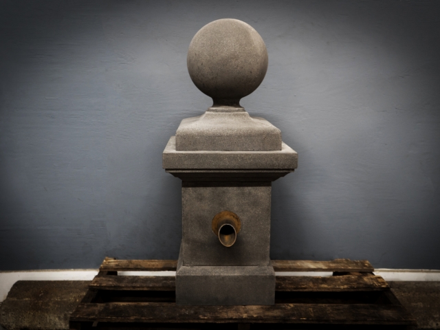 A studio shot of a tall, gray, stone pedestal fountain. The pedestal has a square base, a rounded top, and a spherical finial. A brass-colored spout opening is visible on the front of the pedestal. It is placed on a rough wooden pallet in front of a dark gray wall.