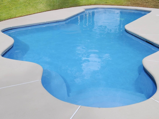 A high-angle view of a clean, irregularly shaped swimming pool with bright blue water. The pool has a white concrete deck surrounding it, with a dark blue tile border at the waterline. The deck and pool have gentle curves and smooth lines.