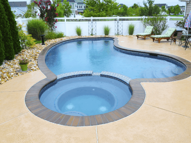 A view of a custom-shaped swimming pool with an integrated circular hot tub. The pool and hot tub are surrounded by a textured, beige concrete patio with a decorative brick border. The hot tub is slightly elevated and has a tiled wall where water can spill into the main pool.