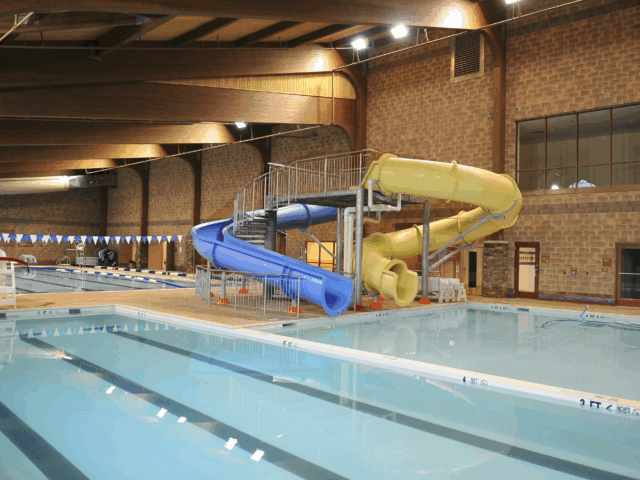 An indoor swimming pool facility featuring a long lap pool and a separate, shallow leisure pool with two twisting water slides. The slides are yellow and blue, and a metal staircase leads up to their entrance.
