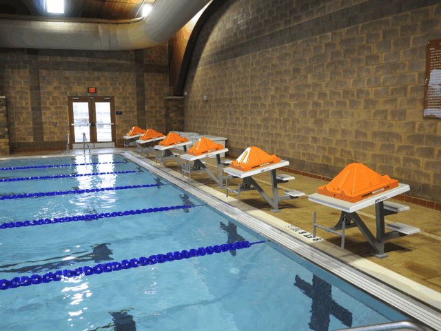 An indoor swimming pool with lap lanes and lane dividers. The pool deck is made of brown tile and a light-colored brick wall. Along the side of the pool, five small diving podiums with bright orange caps are lined up.