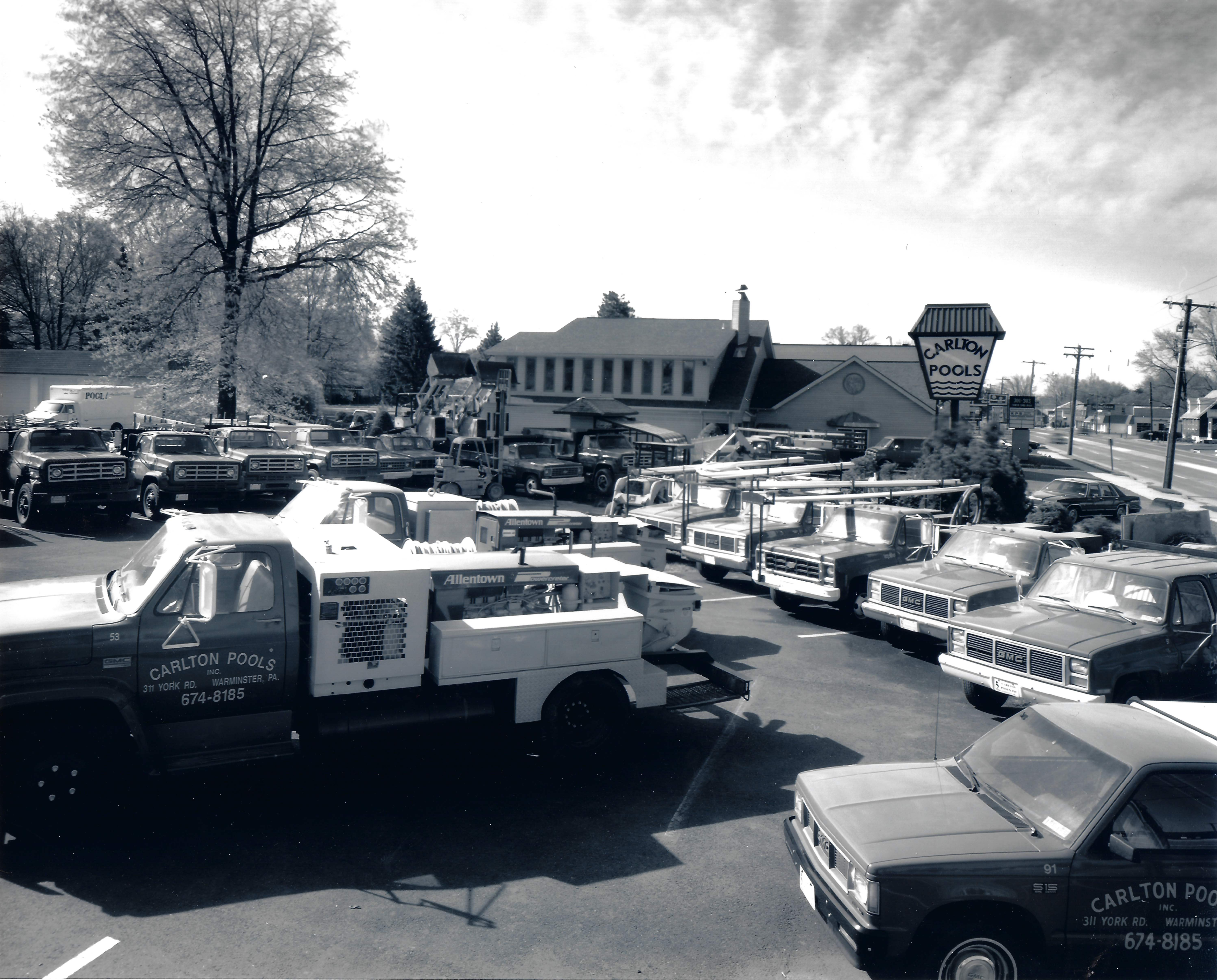 A faded, vintage photograph of a large parking lot filled with red and white trucks, many of which are marked with "CARLTON POOLS". The trucks are service vehicles, some with ladder racks and equipment boxes. In the background, there is a building with a tall sign that also reads "CARLTON POOLS".