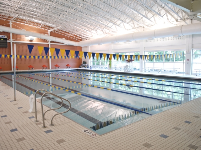 An indoor lap pool at the Somerset Hills YMCA. The pool has swim lanes marked with blue and yellow lane dividers. Yellow and blue pennant flags hang from the ceiling across the width of the pool. A scoreboard is visible on the far wall. The pool deck is tiled.