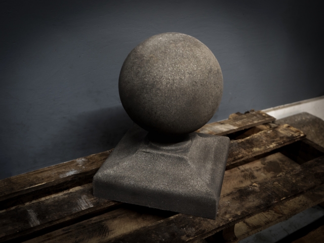 A studio shot of a dark gray, spherical stone finial sitting on a square base. The finial has a coarse, granular texture. It is placed on a rough wooden pallet in front of a dark blue-gray background.
