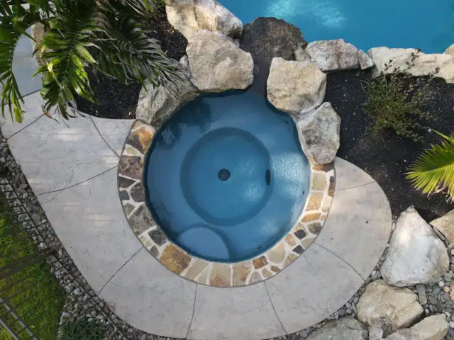 A top-down aerial view of a circular inground spa surrounded by custom stone and concrete decking, integrated into a larger pool and landscaping area.