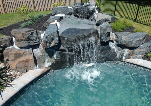 DCIM/977MEDIA/DJI_1011.JPG A natural rock waterfall feature constructed from large, dark, rugged boulders, cascading into a blue swimming pool, framed by a black metal fence.