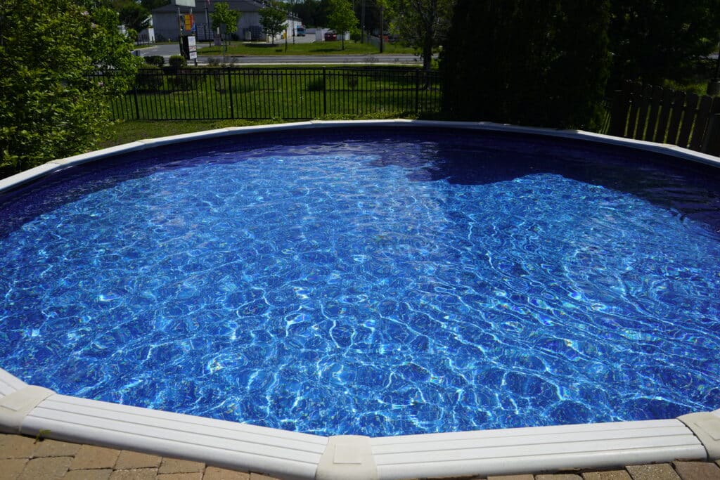 This image features a vibrant, circular above-ground swimming pool filled with clear, shimmering blue water. The pool is framed by a clean white top rail and is situated adjacent to a light-colored brick paver patio in the foreground.

In the background, a black metal fence separates the pool area from a lush green lawn, several leafy trees, and a street with commercial buildings in the distance under a clear, bright sky.