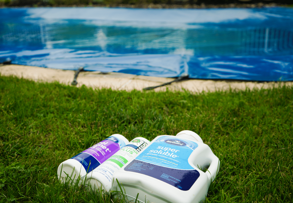 A low-angle, close-up shot focuses on three white plastic pool chemical containers sitting on a green lawn. The largest container in the foreground is labeled "super soluble" granular chlorinator, while two smaller bottles, labeled for metal prevention and algaecide, lie behind it. In the background, a bright blue swimming pool cover is stretched over an inground pool, secured to the surrounding stone deck with black straps. The background is slightly out of focus, emphasizing the maintenance products in the grass.