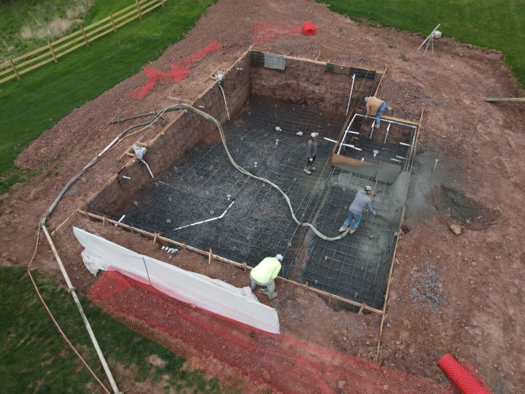 An aerial, high-angle shot captures a construction crew in the process of pouring a concrete pool floor. Three workers stand inside a large, rectangular excavated pit lined with a dense grid of steel rebar and plumbing pipes. One worker directs a thick hose discharging wet concrete onto a portion of the floor, while another, wearing a bright neon green shirt, stands near the edge. The construction site is surrounded by exposed reddish-brown dirt, patches of green grass, a wooden fence, and rolls of orange safety netting.
