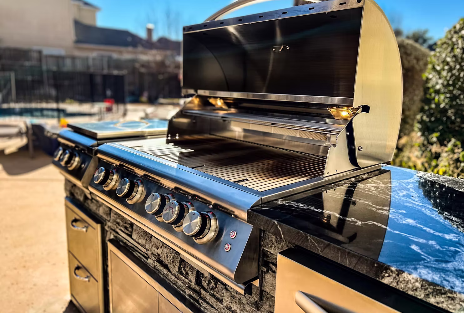 A close-up, low-angle shot of a premium outdoor stainless steel grill built into a stone veneer island with a polished black marble countertop. The grill hood is open to reveal the cooking grates and internal lighting, and the front panel features multiple brushed-metal control knobs, set against a sunny, blurred outdoor patio background.