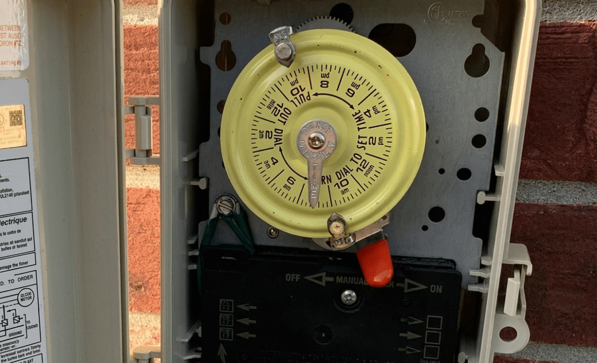 A Close-up of a vintage Intermatic mechanical pool timer mounted inside a beige weatherproof box on a red brick wall. The central yellow 24-hour dial features black numbers and silver metal "on/off" trippers. Below the dial is a black control panel with a red manual override toggle switch and technical diagrams.