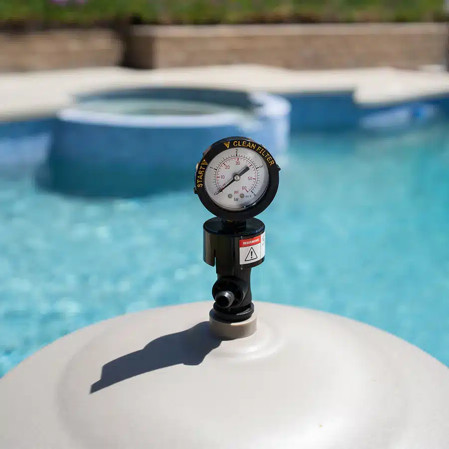 A close-up of a swimming pool filter pressure gauge showing a reading of approximately 18 psi, with a bright blue backyard pool and hot tub blurred in the background.