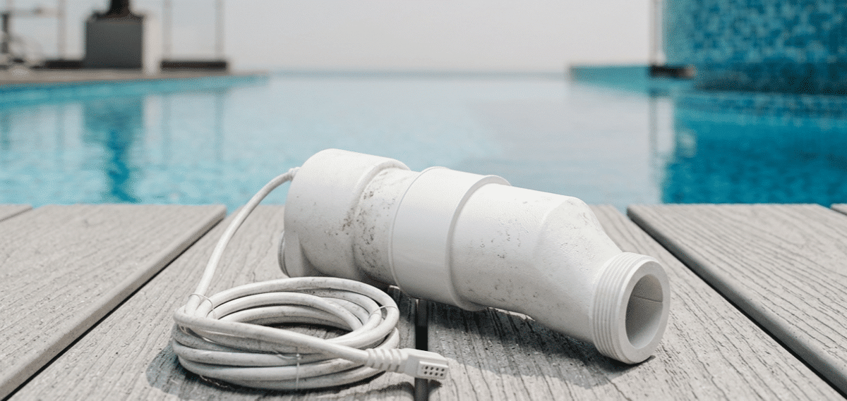 A photograph of a white, unbranded salt cell pool chlorinator resting on a light gray composite or stone deck. The device shows realistic weathering and a matte texture, with its white power cable neatly coiled beside it.